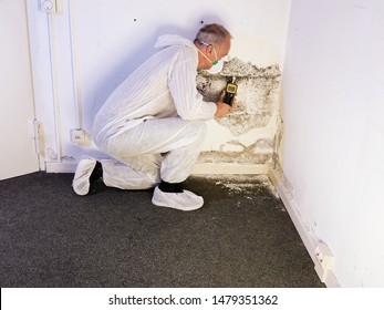 a professional pest control contractor or exterminator in his protection work wear for mold pests and chemicals kneeling at a mold destroyed wall with a moisture meter and check the humidity