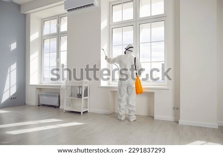 Professional pest control inside the house. Exterminator wearing protective suit, mask and gas respirator spraying insecticide from yellow sprayer bottle over white window frame in modern room at home