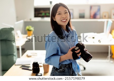 Professional Photographer. Portrait Of Smiling Asian Woman Holding Equipment And Looking Smiling To Camera Standing At Home Office. Lady Taking Photos Posing With Photocamera. Photography Concept