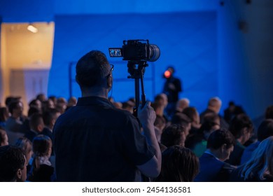 Professional photographer records a corporate event using a tripod and camera. The audience, focused and engaged, listens to a speaker under blue lighting. - Powered by Shutterstock