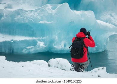 Professional photographer in red coat and black backpack taking picture of glacier ice