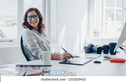 Professional photographer sitting at her office desk looking away and smiling. Woman in office with digital graphic tablet and drawing pen. - Powered by Shutterstock