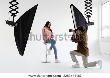 Professional photographer taking picture of young woman in casual wear, having photoshoot in modern studio with lighting equipment on white background