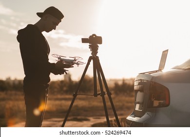Professional photographer working outdoor at sunset. Silhouette of young man standing with drone and camera on tripod. Hobby, leisure, work concept