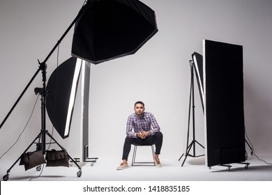 Professional photography studio showing behind the scenes lights. fashion handsome young man model at studio in the light flashes, sitting and looking at camera. indoor studio shot on grey background.