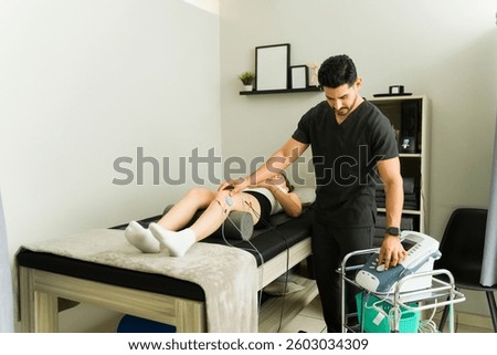Professional physiotherapist setting up electrotherapy device, applying electrodes to patient's leg during medical treatment in clinical setting