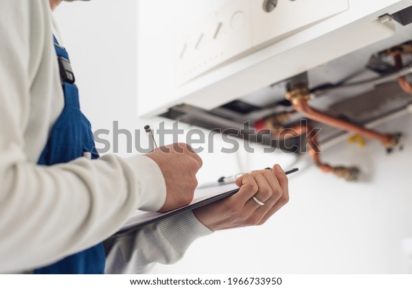 Professional plumber checking a boiler at home\
and writing on a\
clipboard
