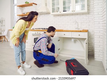 Professional plumber fixing problems with the sink drain in a modern white kitchen in a young lady's house. Plumber crouching on the floor and thinking of how to unclog the clogged plastic sink pipe