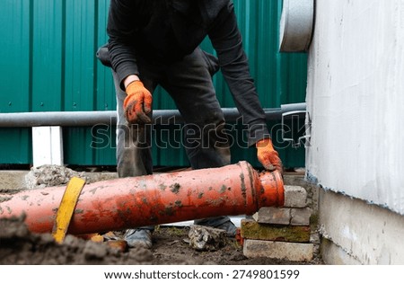 Professional plumber installing a large orange PVC sewage pipe during house renovation. Construction worker in gloves connecting drainage system in the backyard.