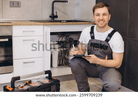 Professional plumber taking notes during inspection of pipes under kitchen sink, ensuring efficient maintenance and repairs