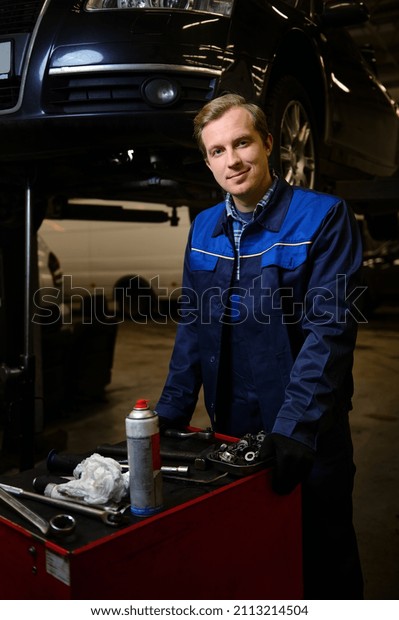 Professional portrait of a handsome Caucasian man,\
auto mechanic, technician, car engineer standing near a box with\
set of tools for repairing a lifted automobile on a hoist in the\
repair shop garage