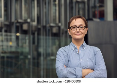 Professional Portrait Of Middle - Aged Older Woman In Her Forties - Fifties,  Wearing Glasses And Shirt. Headshot Of A Factory Business Unit Manager. Blurred Industrial Background.