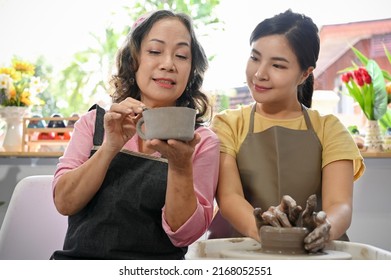 A Professional Potter Teaching And Coaching Her Young Female Student Making A Pottery In The Workshop. Charming Asian Woman Moulding Clay On Potter's Wheel.