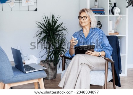 Professional Psychotherapy online. Middle aged female Psychologist having video call consultation on laptop while sitting on armchair In office. Specialist talking, counseling, helping patient online