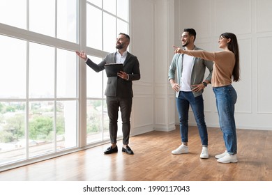 Professional Real Estate Agent Showing Spacious New Apartment To Young Couple, Looking And Pointing At Beautiful View From Big Panoramic Picture Windows. Happy Man And Woman Ready To Become Homeowners