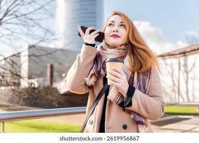 Professional redhead professional pausing during workday, listening to voice message while holding coffee, standing near contemporary urban office building under bright sunlight