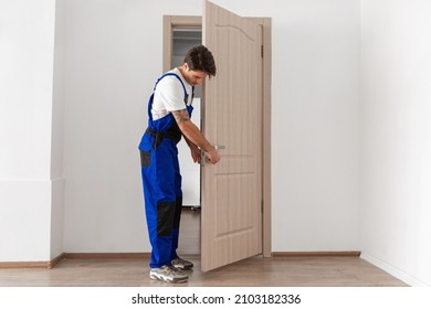 Professional Repair Service. Full Body Legth Of Young Locksmith In Blue Uniform Standing In Doorway Installing Doorknob Handle. Installation Of Lock Or Knob On Front Door Entrance. Maintenance Concept