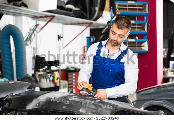 Professional repairman in car workshop sanding\
automobile bumper, preparing for\
painting
