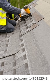 Professional Roofer Worker In Uniform Work Wear Using Air Or Pneumatic Nail Gun And Installing Asphalt Or Bitumen Shingle On Top Of The New Roof Under Construction Residential Building