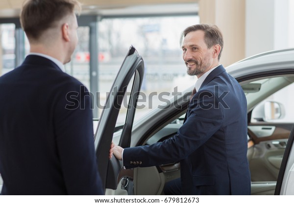 Professional salesman holding car door for his\
client. Businessman getting into his newly bought car at the\
dealership salon career job occupation customer consumer service\
helping assisting\
showing