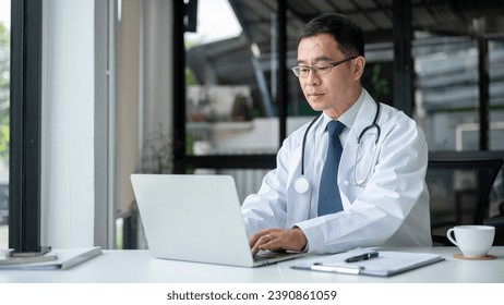 A professional senior Asian male doctor is working on his laptop computer in his office at the clinic, reading medical cases online or responding to emails. - Powered by Shutterstock