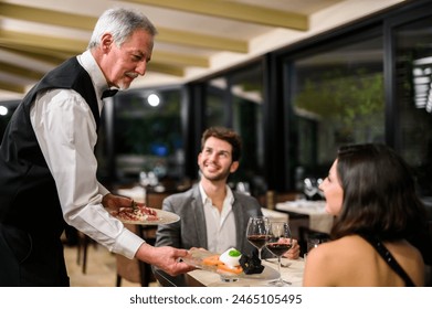 Professional senior male waiter presents a dish to a smiling young couple during a romantic dinner - Powered by Shutterstock