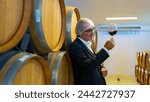 Professional Senior man sommelier tasting and sniffing red wine in wine glass at wine cellar with wooden barrel. Winery brewery liquor wine factory manufacturing industry and winemaker concept. 