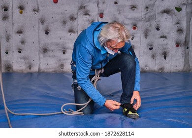 Professional Senior Man Wearing Protective Face Mask Tying His Climbing Shoes Getting Ready To Climb On An Artificial Rock Climbing Wall. Extreme Sports Concept.