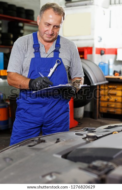 Professional serious auto mechanic recording\
list of works on car repair in auto workshop\
