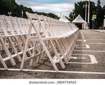 Professional setup of rows of white folding chairs for an outdoor ceremony or event. Clean, minimalist furniture arrangement in a public square against a backdrop of trees and white marquees.