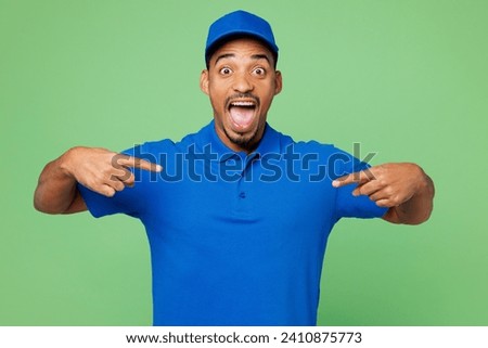 Professional shocked happy delivery guy employee man wear blue cap t-shirt uniform workwear work as dealer courier point index fingers on himself isolated on plain green background. Service concept