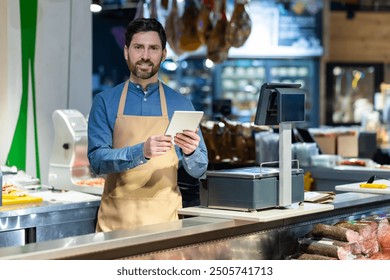 Professional shopkeeper in apron using tablet at delicatessen counter of modern grocery store. Warm smile conveys friendly service. Background shows variety of products - Powered by Shutterstock