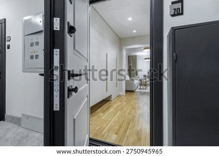 A professional shot looking through an open black entrance door into a bright modern apartment. The view reveals a minimalist hallway with white built-in wardrobes, warm wooden flooring, and a glimpse of a contemporary living room in the background.