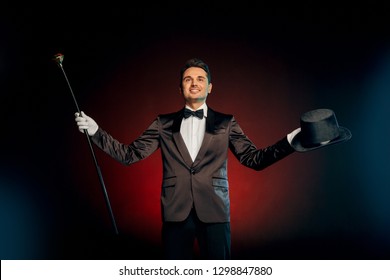 Professional showman wearing suit standing isolated on black and red background holding top hat and pimp cane starting show smiling confident