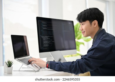 Professional and smart young Asian male programmer coding at his desk, working on his project, using laptop and computer. - Shutterstock ID 2246595355