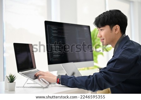 Professional and smart young Asian male programmer coding at his desk, working on his project, using laptop and computer.