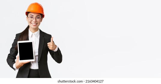 Professional Smiling Asian Female Construction Manager, Factory Engineer In Business Suit And Safety Helmet Showing Thumbs-up And Digital Tablet Screen With Positive Graph, White Background