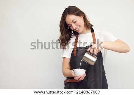 Professional smiling barista woman in studio serving coffee, pour milk from a jug in a coffee cup. Café latte. Isolated on white background.