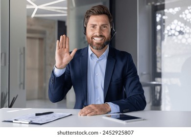 Professional smiling businessman in suit engaged in a video call, waving at the camera with a friendly gesture, in a bright modern office setting. - Powered by Shutterstock