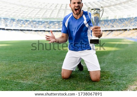 professional soccer player in blue and white uniform with sports cup standing on knees on football pitch and shouting at stadium