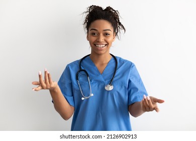 Professional Specialist. Smiling Black Female Doctor Wearing Blue Medical Uniform Coat And Stethoscope Talking To Camera, Explaining And Gesturing, Standing Isolated Over White Studio Background Wall