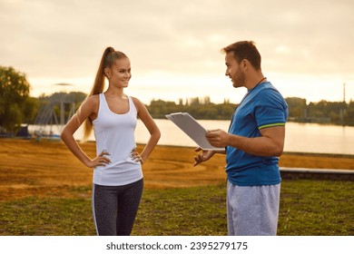 Professional sports trainer talking to young female athlete. Man personal coach with clipboard in hand and happy beautiful slim woman in tank top standing on green lawn and discussing workout plan - Powered by Shutterstock