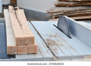 A professional static shot of a circular table saw machine with a sharp saw blade ready for work. Neatly cut wooden beams and planks are stacked on the side. The background shows more lumber in a blurred outdoor or semi-outdoor construction setting in Ukraine. This image highlights precision, carpentry preparation, and industrial equipment. Perfect for DIY guides, hardware catalogs, and woodworking business websites. - Powered by Shutterstock - Get 15% off with code: PIKWIZARD15