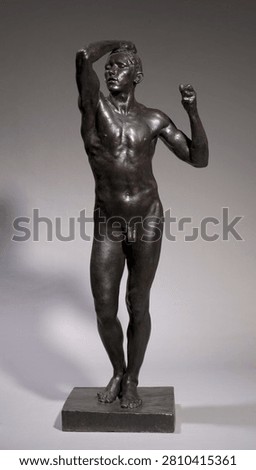 A professional studio photograph captures a dramatic, full-length bronze sculpture of a nude young male figure with refined classical modeling.