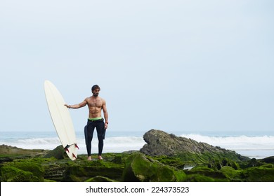 Professional Surfer In Black Wetsuit Standing On Moss Rocks, Sexy Surfer In Wetsuit Holding A Surfboard And Waiting The Waves