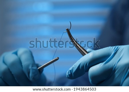 Professional surgeon holding forceps with suture thread on blurred background, closeup. Medical equipment