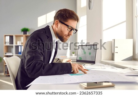 Professional surveyor in suit and glasses sitting at his table in office, working with modern city structure design plans, looking at cadastral maps, and studying plot numbers and boundaries