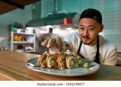 Professional sushi chef wearing protective gloves decorating rolls served on plate at commercial kitchen. Food photography, Asian cuisine, restaurant service concept - Powered by Shutterstock