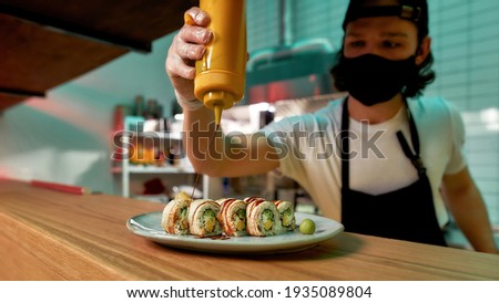 Professional sushi chef wearing protective mask and gloves adding sauce while preparing sushi rolls at commercial kitchen. Food photography, Asian cuisine, restaurant service concept