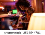 Professional talented multicultural female bass player sitting at music recording studio with bass guitar and recording music. There is a sound technician in a blurry background with channel mixer.